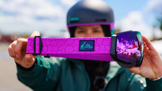 A woman holding the SheJumps designed goggle to show off the purple giraffe patterned goggle strap design. 