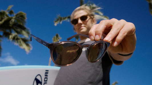 Molly Picklum holding a surfboard under one arm and holding a pair of sunglasses in her other hand. 
