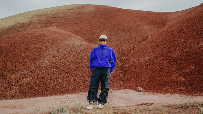 A woman in a bright blue windbreaker and Smith Optics sunglasses stands in an arid landscape.