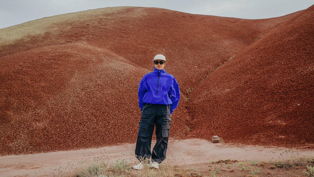 A woman in a bright blue windbreaker and Smith Optics sunglasses stands in an arid landscape.