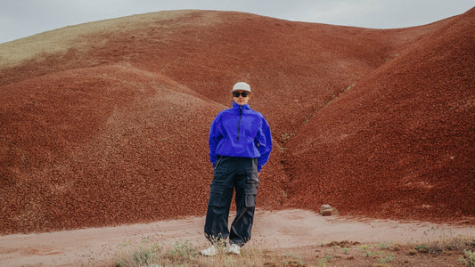 A woman in a bright blue windbreaker and Smith Optics sunglasses stands in an arid landscape.