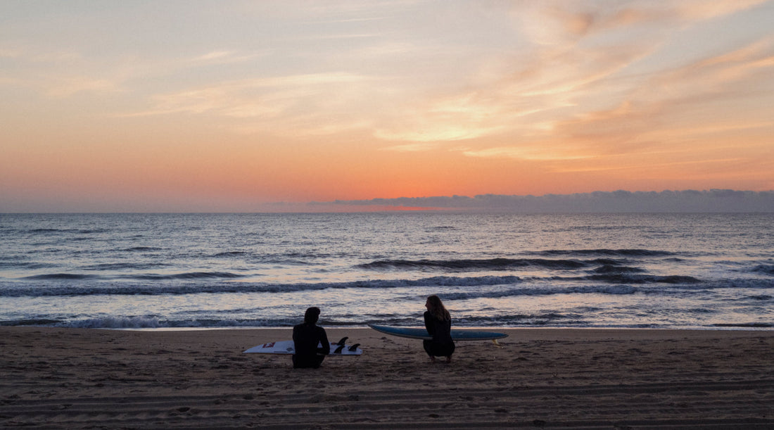 The silhouettes of two people sitting on a beach at sunset.