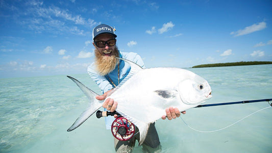 Fisherman wearing Smith sunglasses and hat, standing in the water, exuberantly holding a fish. 