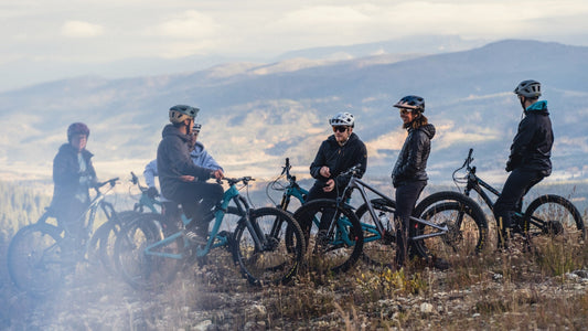 A group of mountain bikers wearing Smith helmets and sunglasses rest on a hill overlooking a valley view.