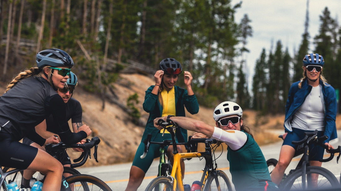 a group of road cyclists wearing Smith helmets and sunglasses, resting by the side of a road in the forest. 
