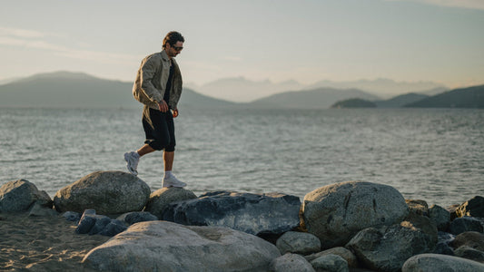 A man walks across rocks at the water's edge with mountains silhouetted in the background behind the water.
