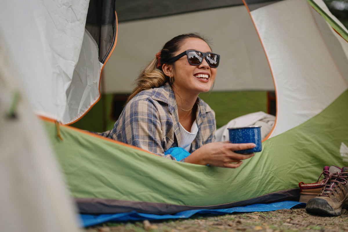 A woman wearing the Smith Lowdown 2 sunglasses smiles while holding a mug while laying in her tent.