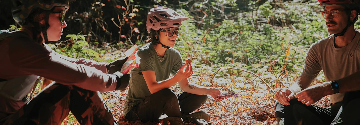 Three people sitting on the ground outdoors, possibly in a forest or natural setting.